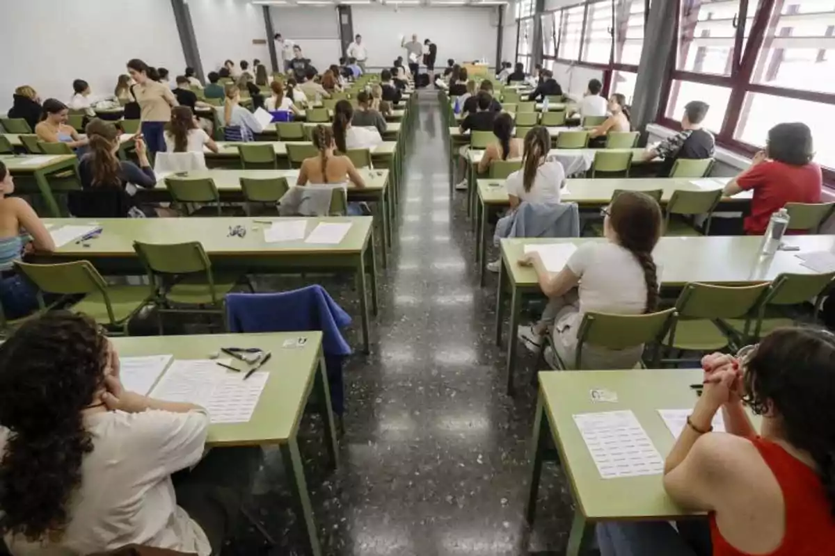 Aula llena de estudiantes sentados en escritorios verdes realizando un examen mientras varios profesores supervisan al fondo Aula llena de estudiantes sentados en escritorios verdes realizando un examen mientras varios profesores supervisan al fondo