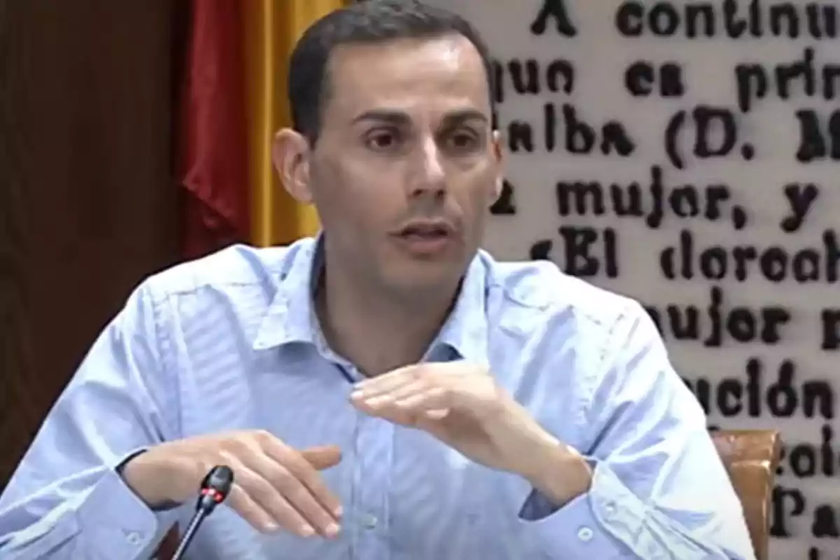 Hombre de cabello corto y camisa azul claro hablando frente a un micrófono con fondo de texto en la pared.