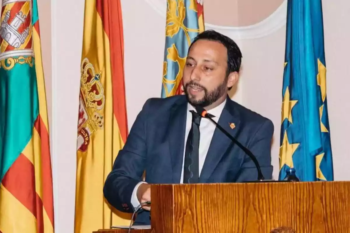 Hombre con barba y traje azul hablando en un podio con varias banderas de fondo