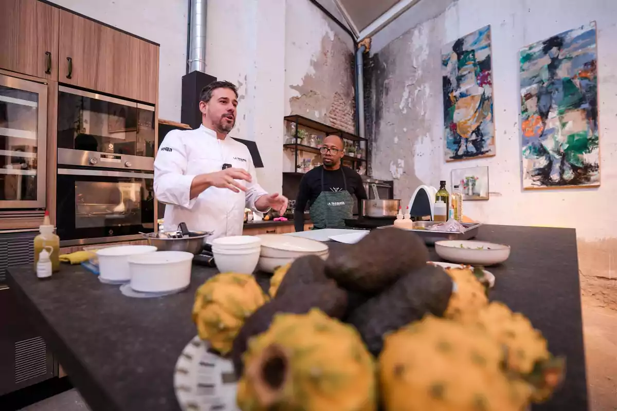 Un chef y un asistente en una cocina moderna con frutas y utensilios en primer plano.