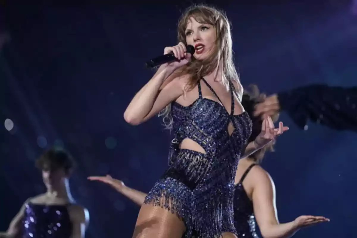 Cantante en el escenario con un micrófono, vestida con un traje brillante y rodeada de bailarines.