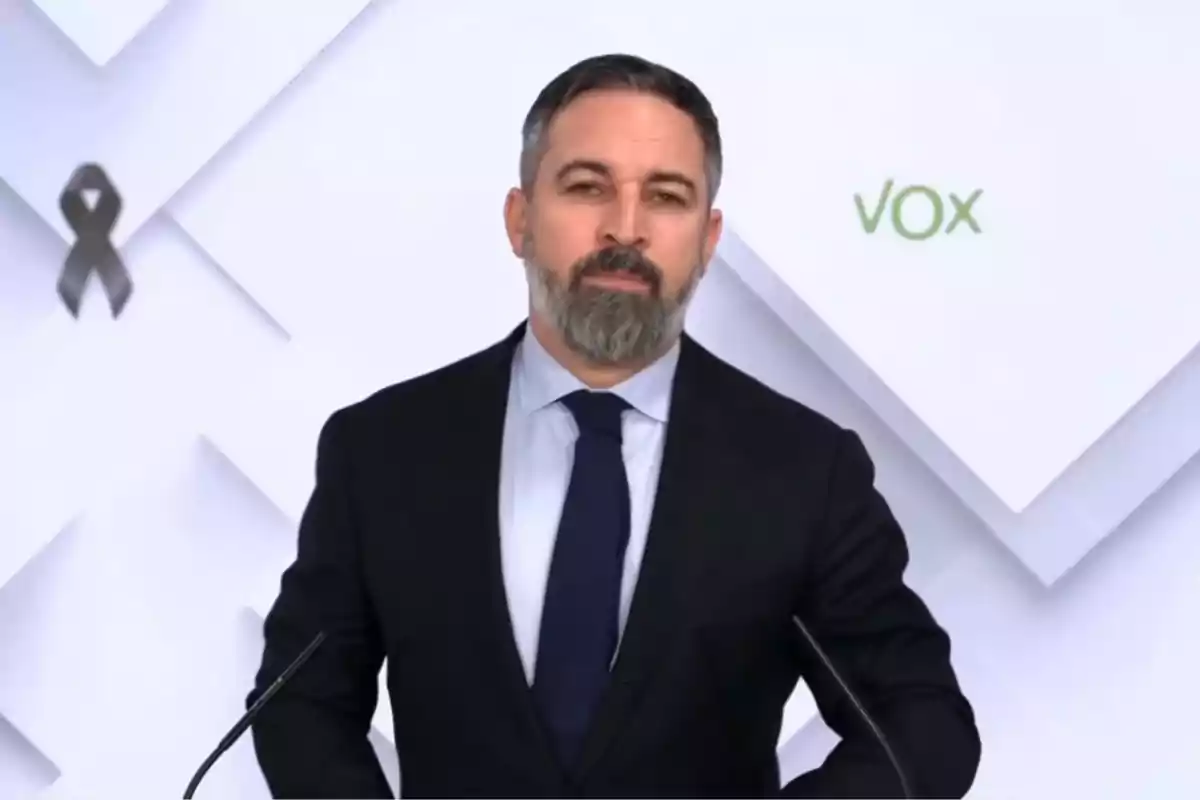 Un hombre con barba y traje oscuro está de pie frente a un fondo blanco con un lazo negro y el logo de Vox.