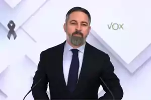 Un hombre con barba y traje oscuro está de pie frente a un fondo blanco con un lazo negro y el logo de Vox.