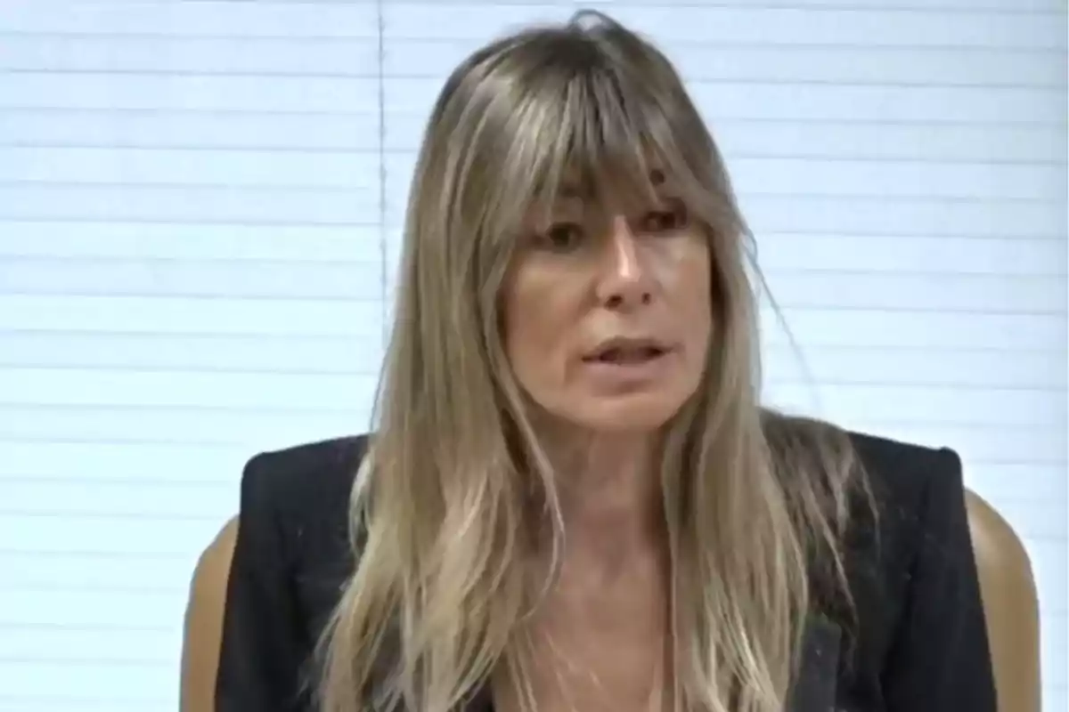 Mujer con cabello rubio y flequillo hablando frente a una persiana blanca.