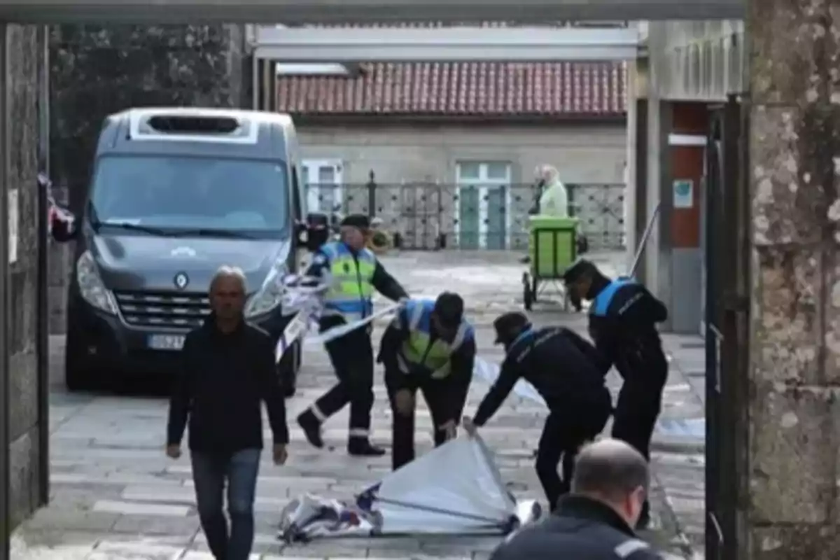 Un grupo de policías levanta una cinta de seguridad en una calle mientras una furgoneta está estacionada cerca y varias personas caminan por la zona.