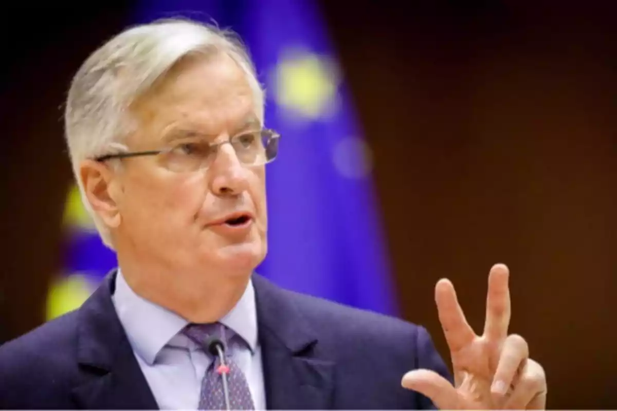 Hombre de cabello canoso con gafas hablando y gesticulando frente a una bandera de la Unión Europea.