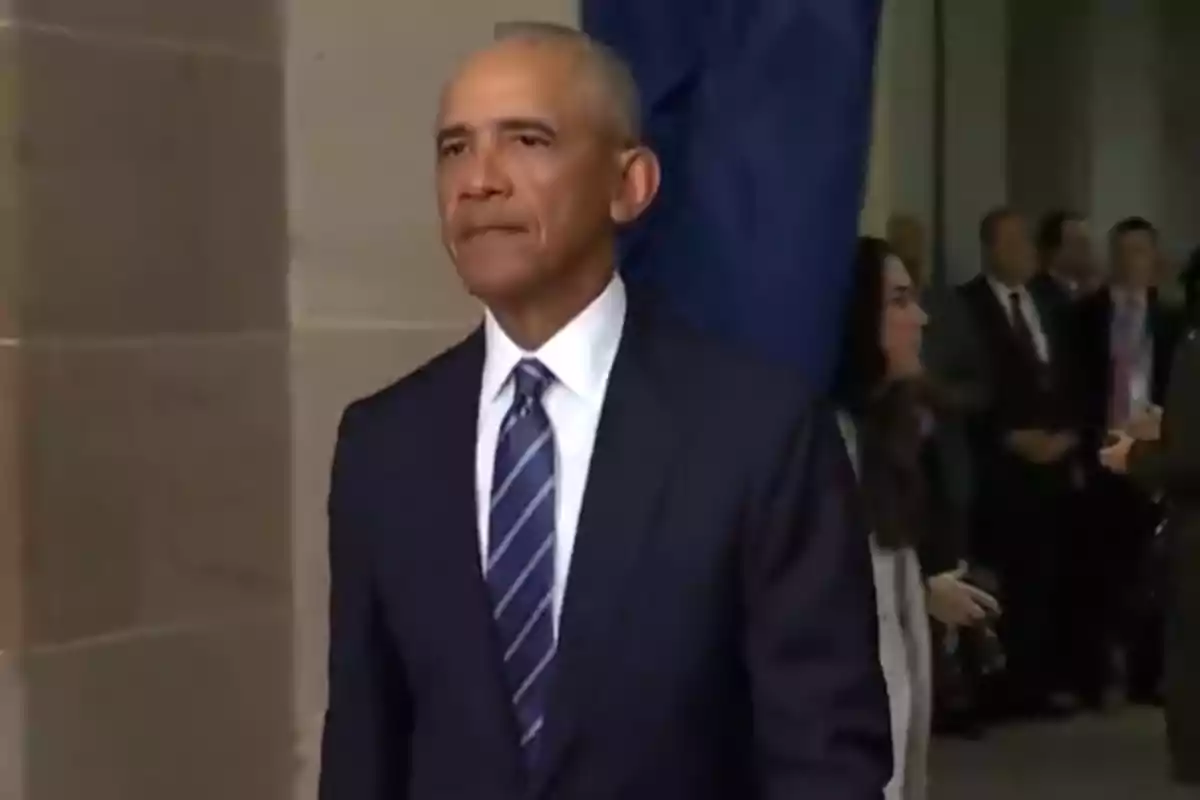 A man in a dark suit and striped tie walks down a hallway with people in the background. A man in a dark suit and striped tie walks down a hallway with people in the background.