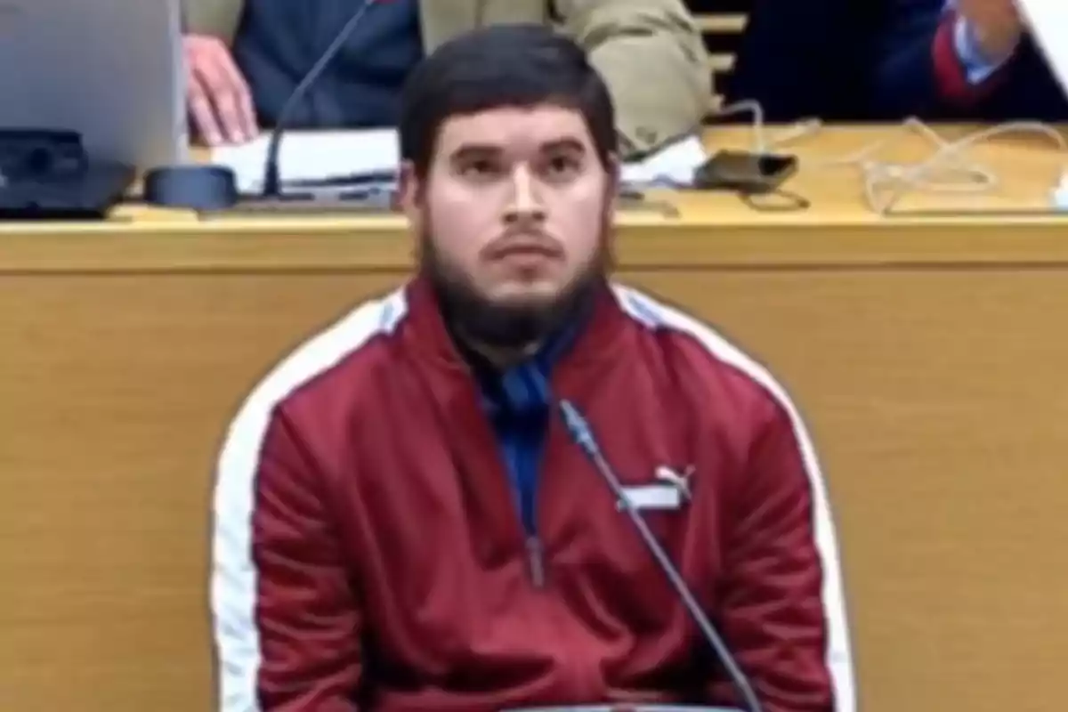 A man in a red jacket is sitting in front of a microphone in a conference room or courtroom.