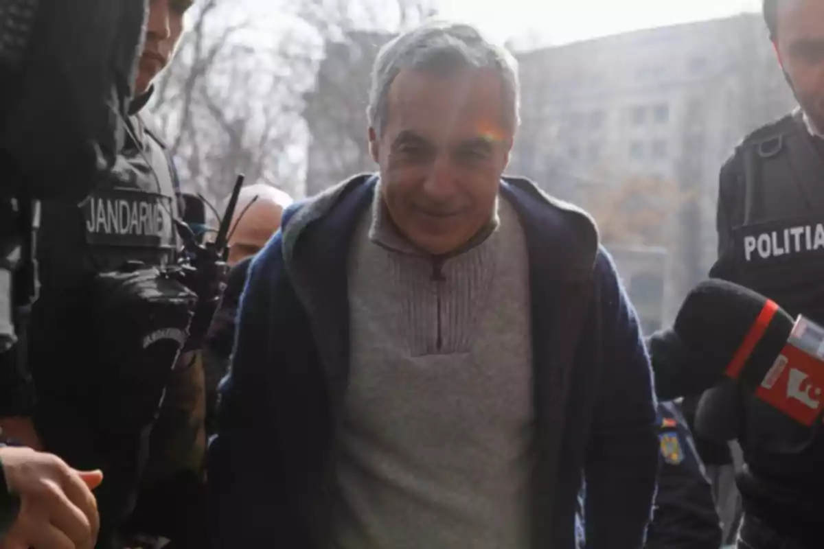 Un hombre de cabello canoso, vestido con un suéter gris y chaqueta azul, rodeado por oficiales de policía y un micrófono de prensa.