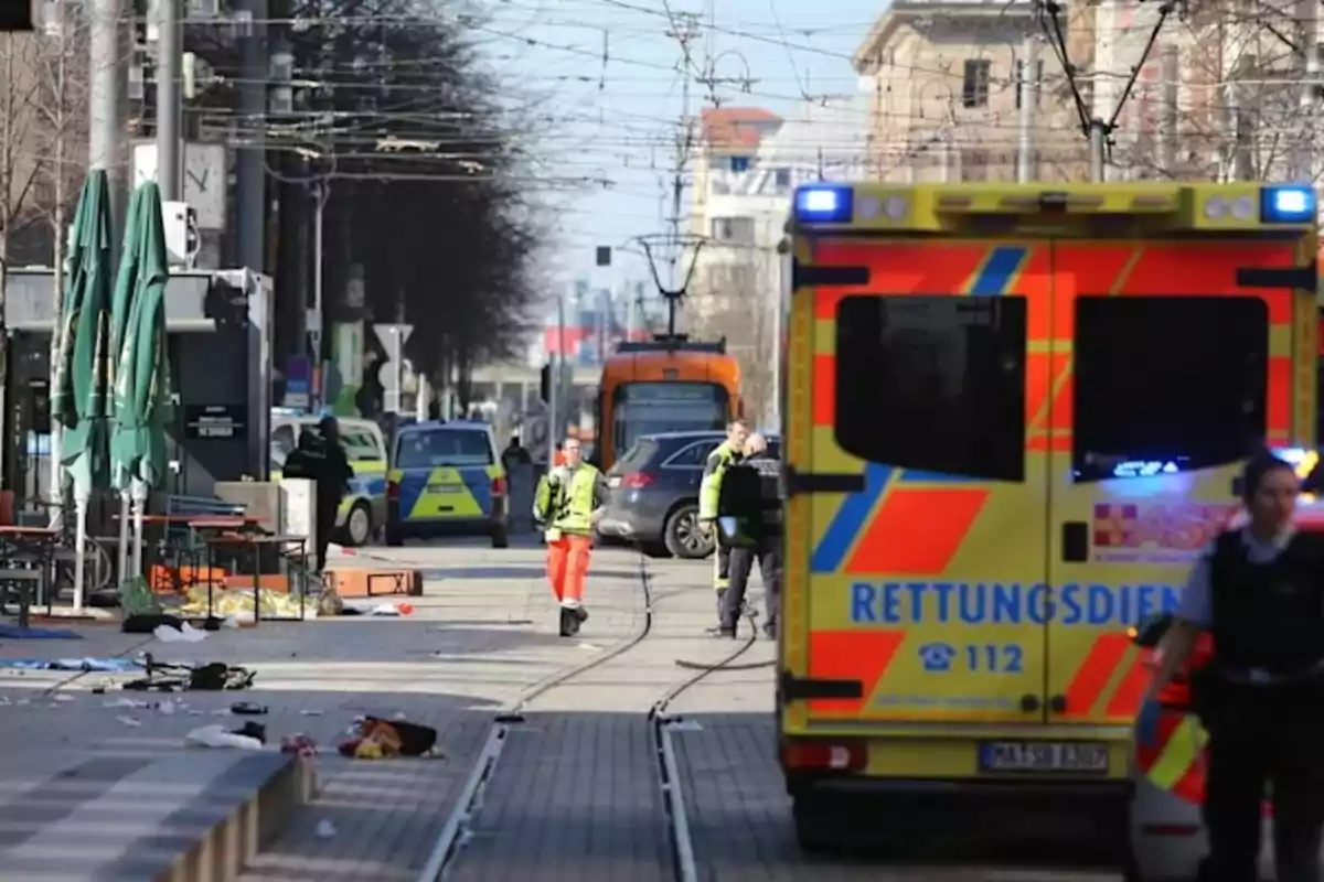 Una escena urbana muestra una ambulancia y varios vehículos de emergencia en una calle con tranvías, mientras personas con chalecos de seguridad caminan por el lugar.