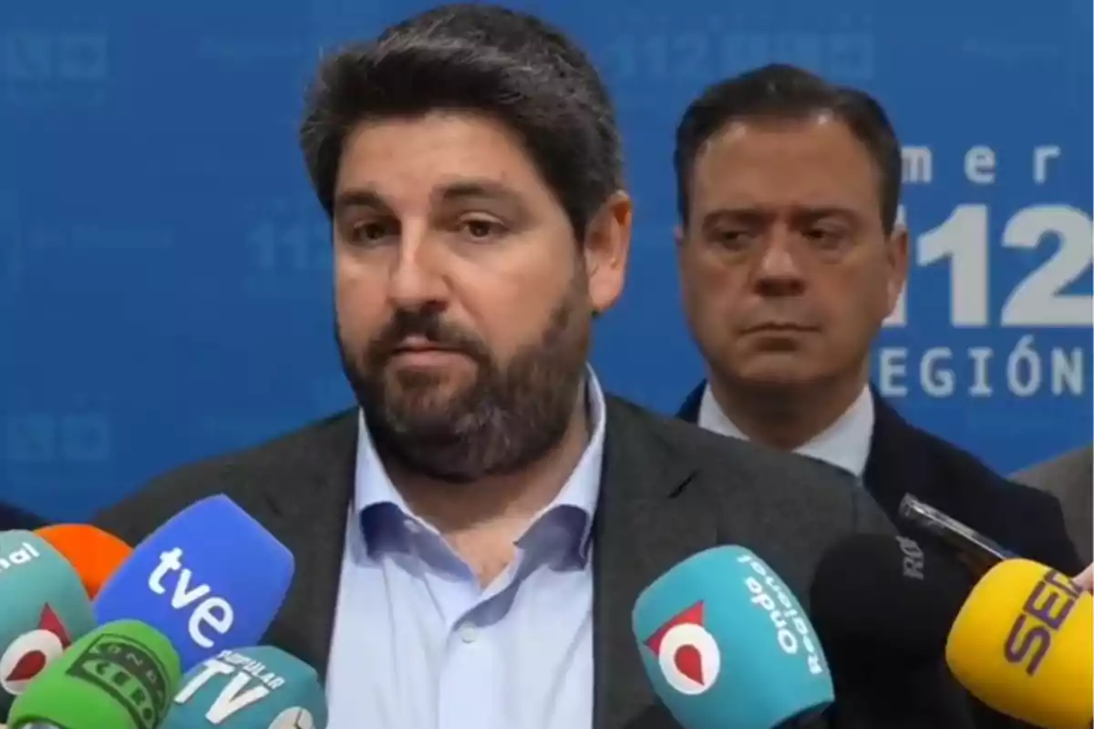 A bearded man speaks in front of several microphones from different media outlets while another man stands behind him.