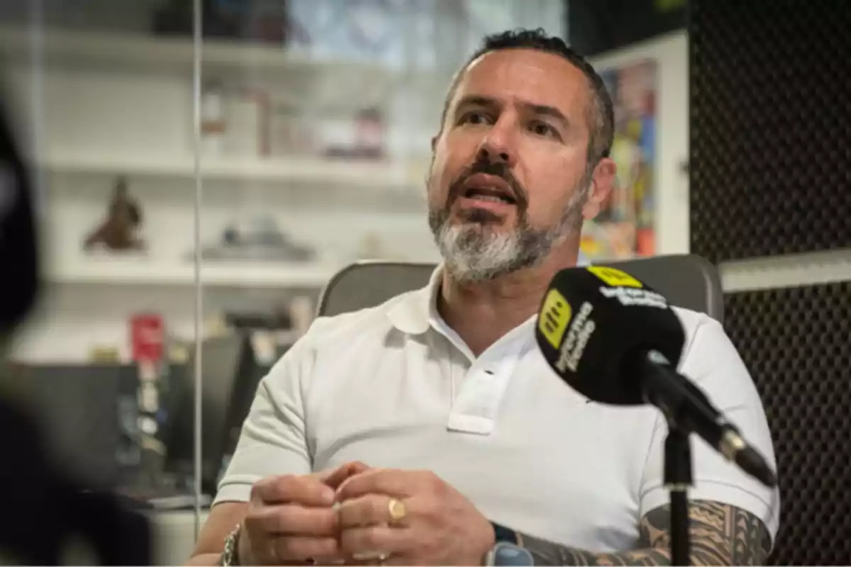 Un hombre con barba y tatuajes en el brazo está hablando en un micrófono durante una entrevista en un estudio. Un hombre con barba y tatuajes en el brazo está hablando en un micrófono durante una entrevista en un estudio.