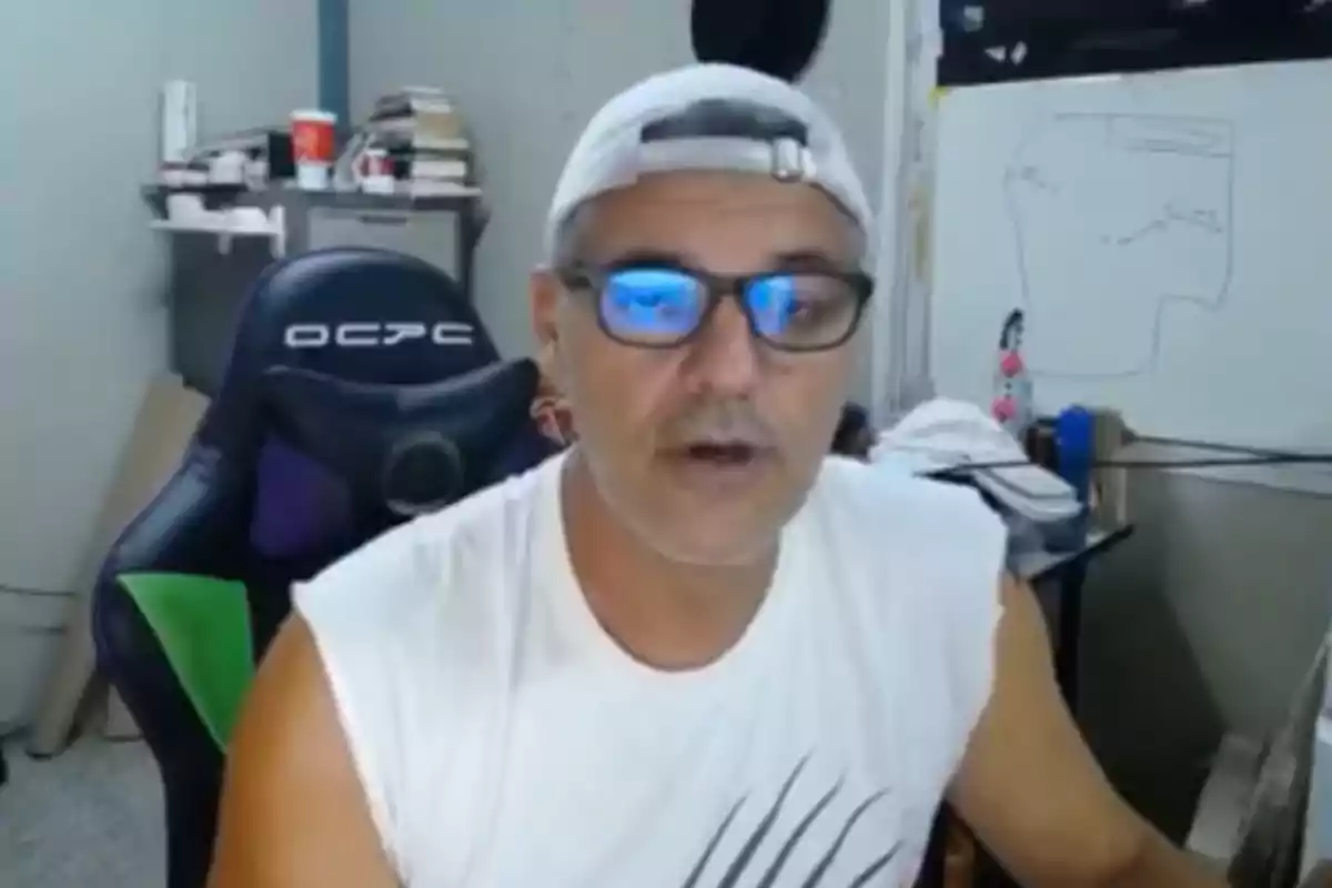 Un hombre con gorra y gafas está sentado frente a una computadora en una habitación con una silla de gaming y una pizarra blanca al fondo.