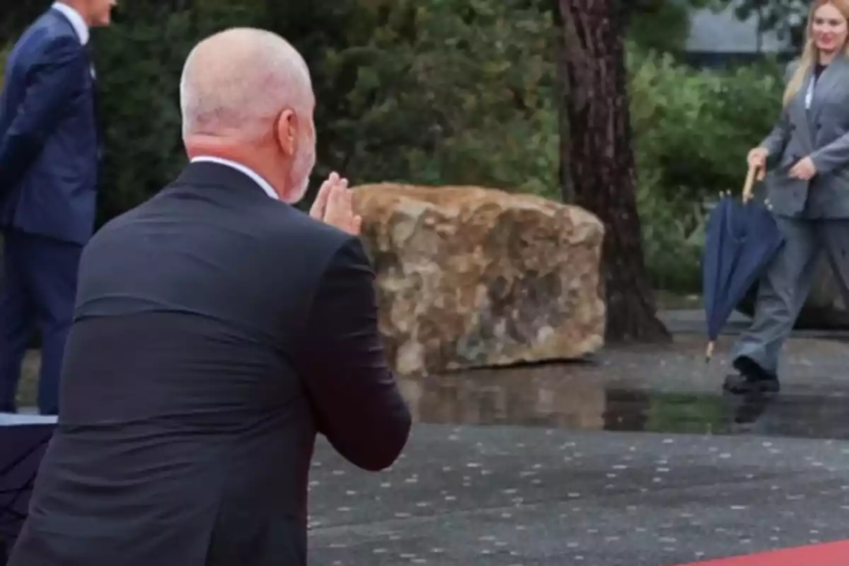 Un hombre de espaldas en traje oscuro parece estar saludando a una mujer que camina hacia él con un paraguas en la mano sobre una alfombra roja en un entorno al aire libre. Un hombre de espaldas en traje oscuro parece estar saludando a una mujer que camina hacia él con un paraguas en la mano sobre una alfombra roja en un entorno al aire libre.