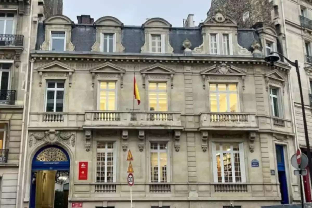 Fachada de un edificio antiguo de piedra con ventanas grandes y una bandera de España en el centro