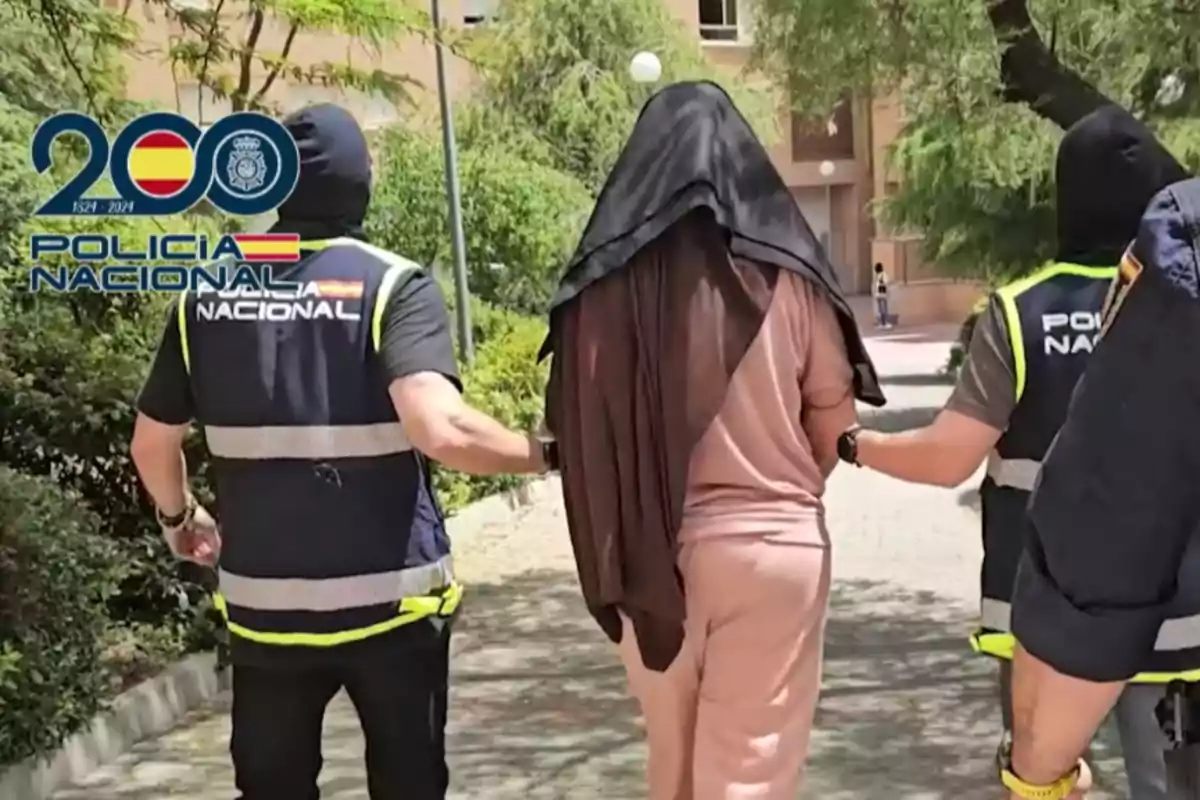 Dos agentes de la Policía Nacional escoltan a una persona detenida con la cabeza cubierta mientras caminan por una zona exterior rodeada de árboles y edificios Dos agentes de la Policía Nacional escoltan a una persona detenida con la cabeza cubierta mientras caminan por una zona exterior rodeada de árboles y edificios