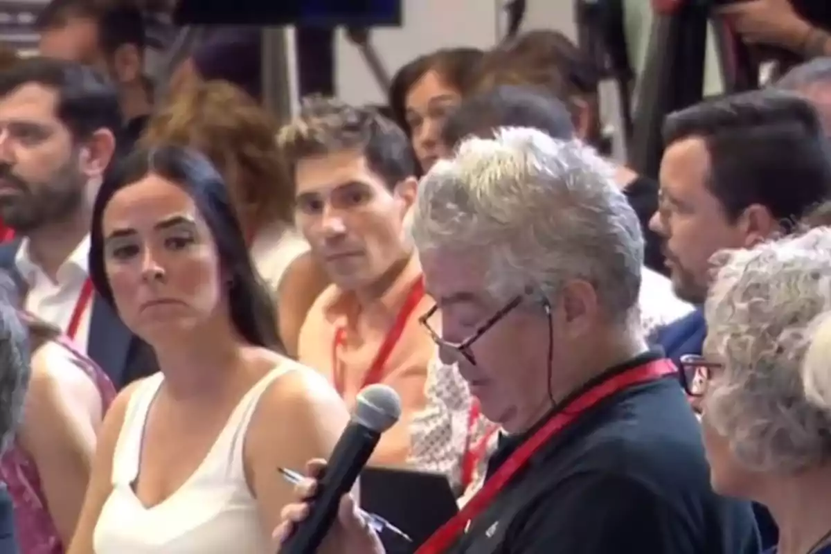 Personas sentadas en una conferencia, con un hombre mayor hablando por micrófono y otros asistentes escuchando atentamente.