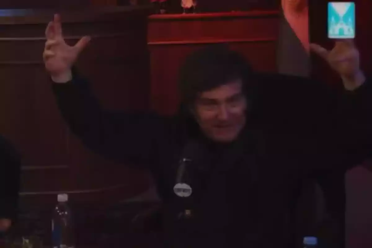 Un hombre con cabello oscuro está sentado frente a un micrófono, levantando ambos brazos en un gesto expresivo, en un ambiente con iluminación tenue y una bandera en el fondo. Un hombre con cabello oscuro está sentado frente a un micrófono, levantando ambos brazos en un gesto expresivo, en un ambiente con iluminación tenue y una bandera en el fondo.