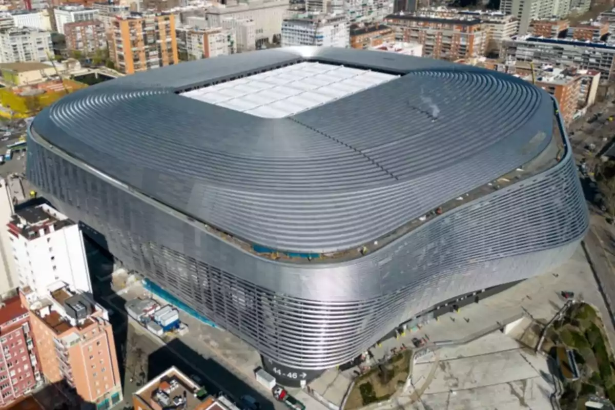 Vista aérea de un estadio moderno con un diseño arquitectónico curvo y una cubierta gris en una zona urbana.