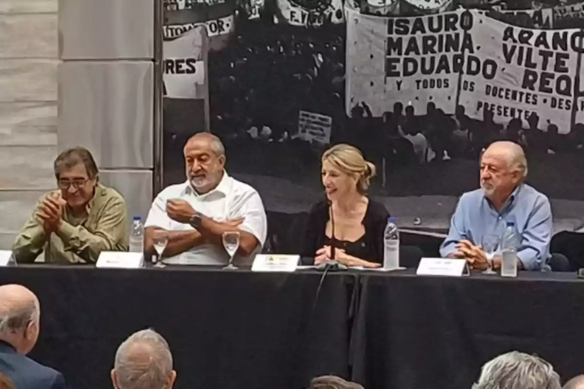 A group of people is sitting at a table during an event or conference, with a background showing banners and a crowd.