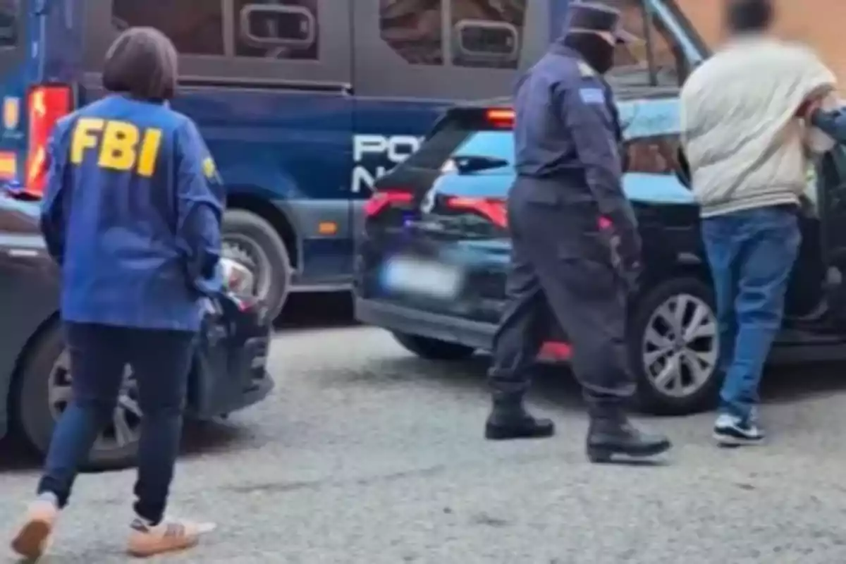 Una persona con chaqueta del FBI y un oficial escoltan a un individuo hacia un vehículo en una calle.