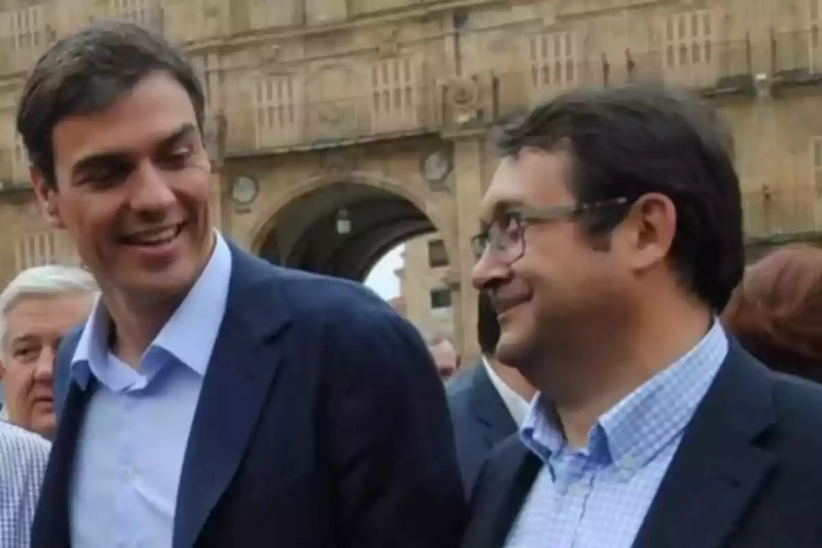 Dos hombres conversando y sonriendo en un entorno urbano con un edificio histórico de fondo.