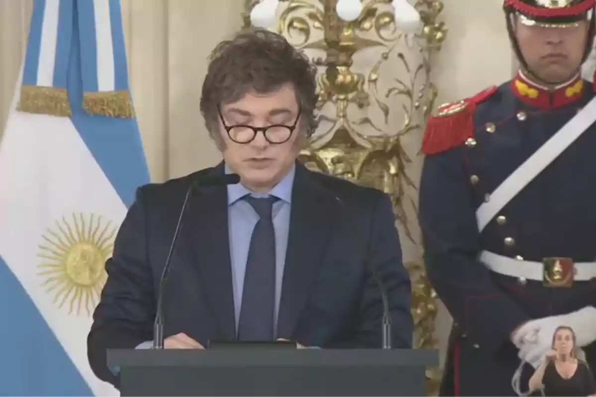 Un hombre con gafas habla en un podio con un micrófono mientras un guardia uniformado está de pie a su lado y una bandera argentina se ve en el fondo.