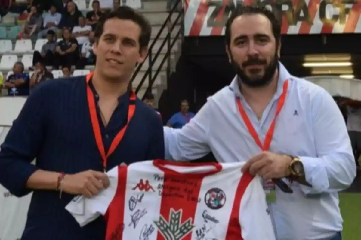 Dos hombres posan en un estadio de fútbol sosteniendo una camiseta firmada del equipo Zamora CF, ambos llevan gafetes rojos y al fondo se ven gradas con público. Dos hombres posan en un estadio de fútbol sosteniendo una camiseta firmada del equipo Zamora CF, ambos llevan gafetes rojos y al fondo se ven gradas con público.