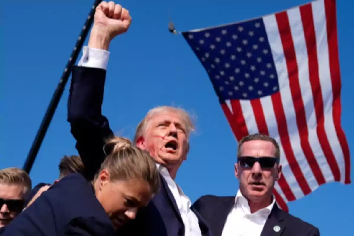 Un hombre levantando el puño rodeado de personas con una bandera de Estados Unidos de fondo. Un hombre levantando el puño rodeado de personas con una bandera de Estados Unidos de fondo.