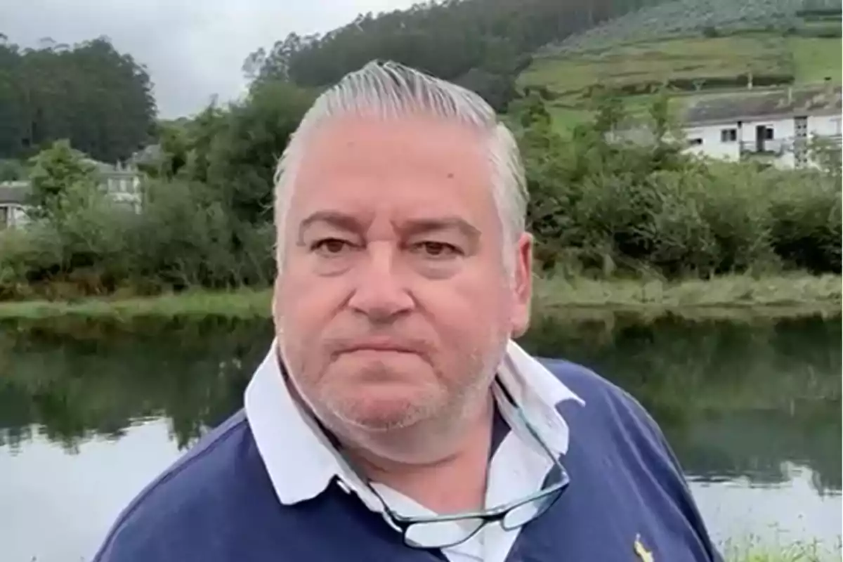 Hombre de cabello canoso y barba incipiente con gafas colgando de su cuello, vistiendo una camisa polo azul, posando frente a un paisaje natural con un río y vegetación al fondo.