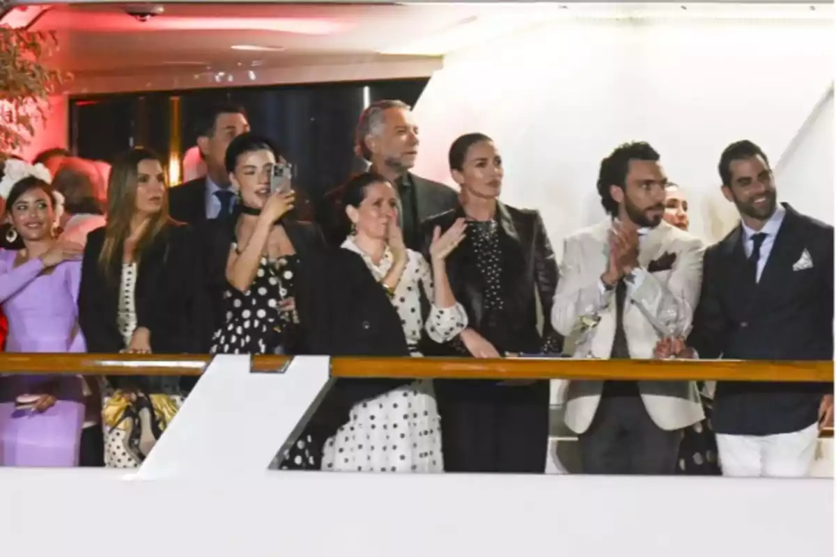 Un grupo de personas elegantemente vestidas se encuentra en un balcón, algunas aplauden mientras otras observan y toman fotos.