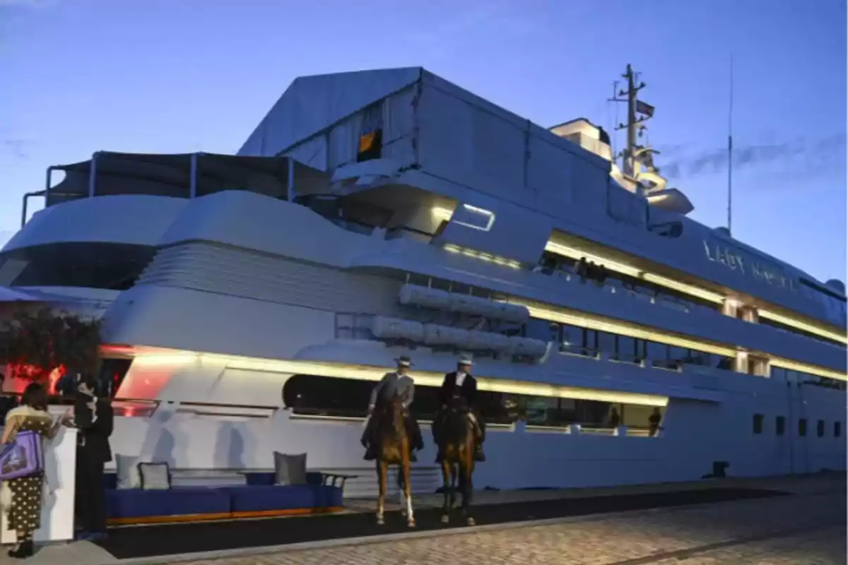 Un yate grande iluminado al anochecer con personas y dos caballos en el muelle. Un yate grande iluminado al anochecer con personas y dos caballos en el muelle.