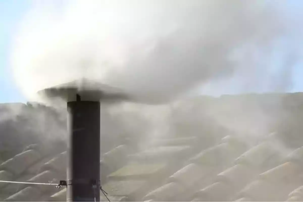 Una chimenea emitiendo humo blanco sobre un fondo de cielo azul y un tejado.
