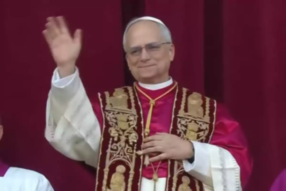 Un hombre vestido con vestimenta religiosa, incluyendo una sotana roja y una estola bordada, levanta la mano en señal de saludo frente a un fondo rojo.