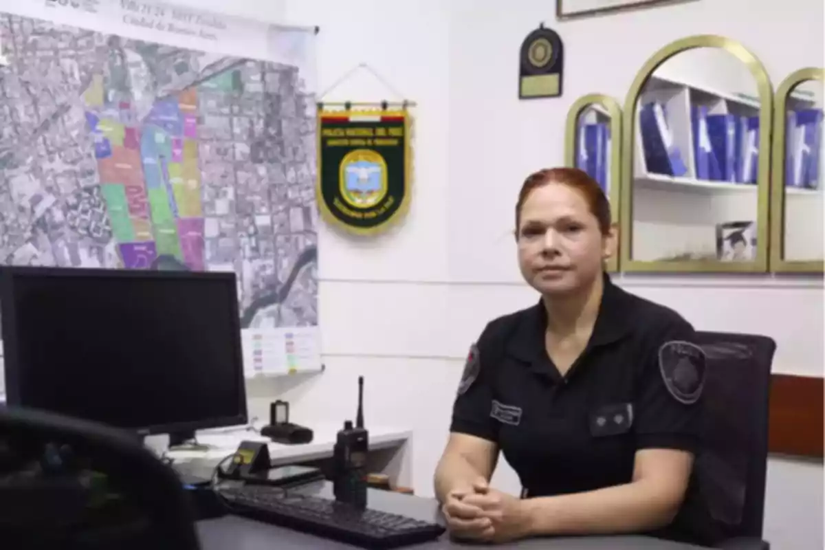 Una mujer con uniforme de policía está sentada en una oficina frente a un escritorio que tiene una computadora y un radio, con un mapa y emblemas en la pared detrás de ella.