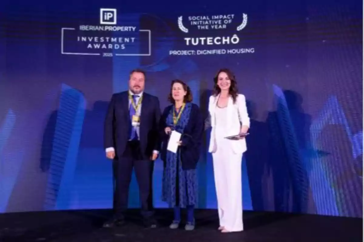 Tres personas posan en un escenario durante un evento de premiación con un fondo que muestra el logotipo de los Iberian Property Investment Awards 2023 y el reconocimiento a una iniciativa de impacto social.
