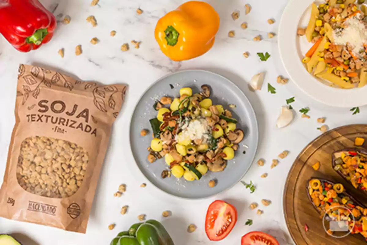 Vista superior de una mesa con platos de comida vegetariana, vegetales frescos, una bolsa de soja texturizada y trozos de tomate, pimiento y ajo sobre una superficie blanca Vista superior de una mesa con platos de comida vegetariana, vegetales frescos, una bolsa de soja texturizada y trozos de tomate, pimiento y ajo sobre una superficie blanca