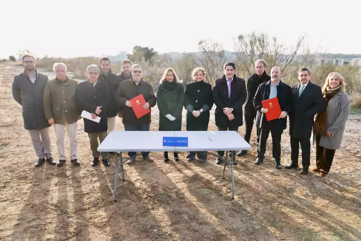 Un grupo de personas posando al aire libre detrás de una mesa con un cartel que dice "Madrid" y algunos sostienen carpetas rojas.