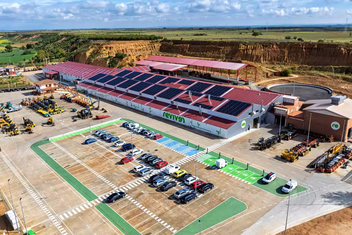 Vista aérea de un complejo industrial con paneles solares en el techo, un estacionamiento lleno de vehículos y maquinaria pesada en el área de trabajo, rodeado de un paisaje natural.