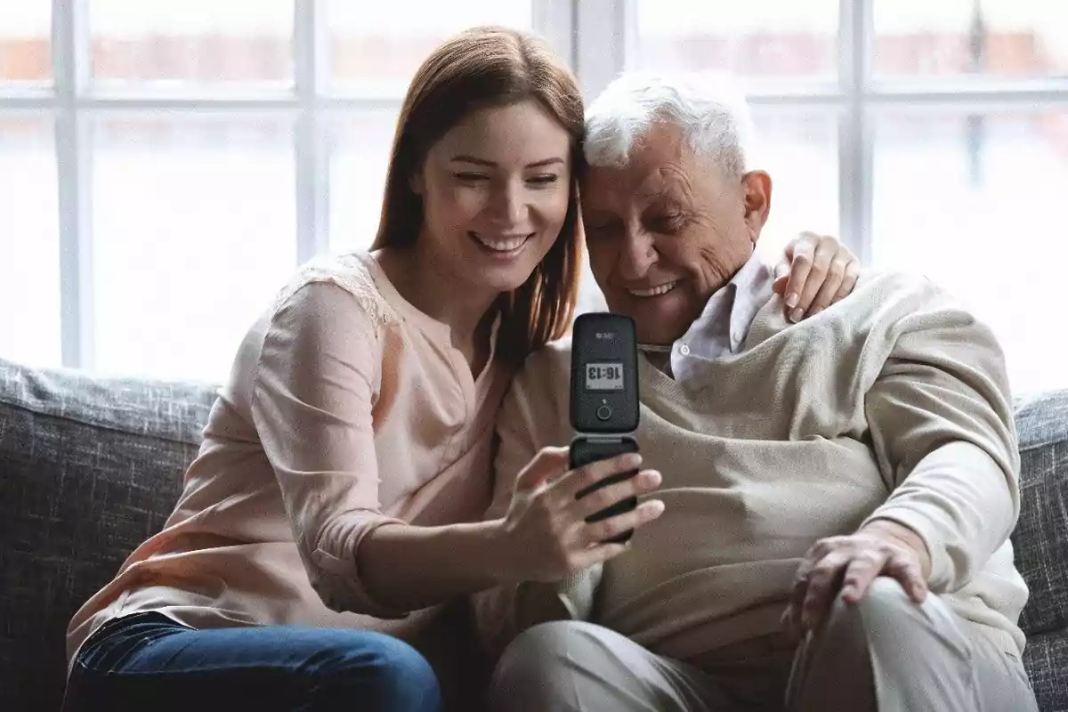 Una mujer joven y un hombre mayor sonríen mientras miran un teléfono plegable juntos en un sofá.