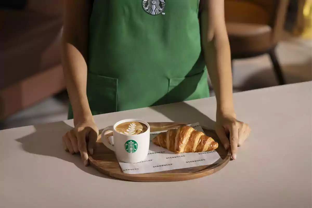 Una persona con un delantal verde de Starbucks sostiene una bandeja con un café y un croissant.