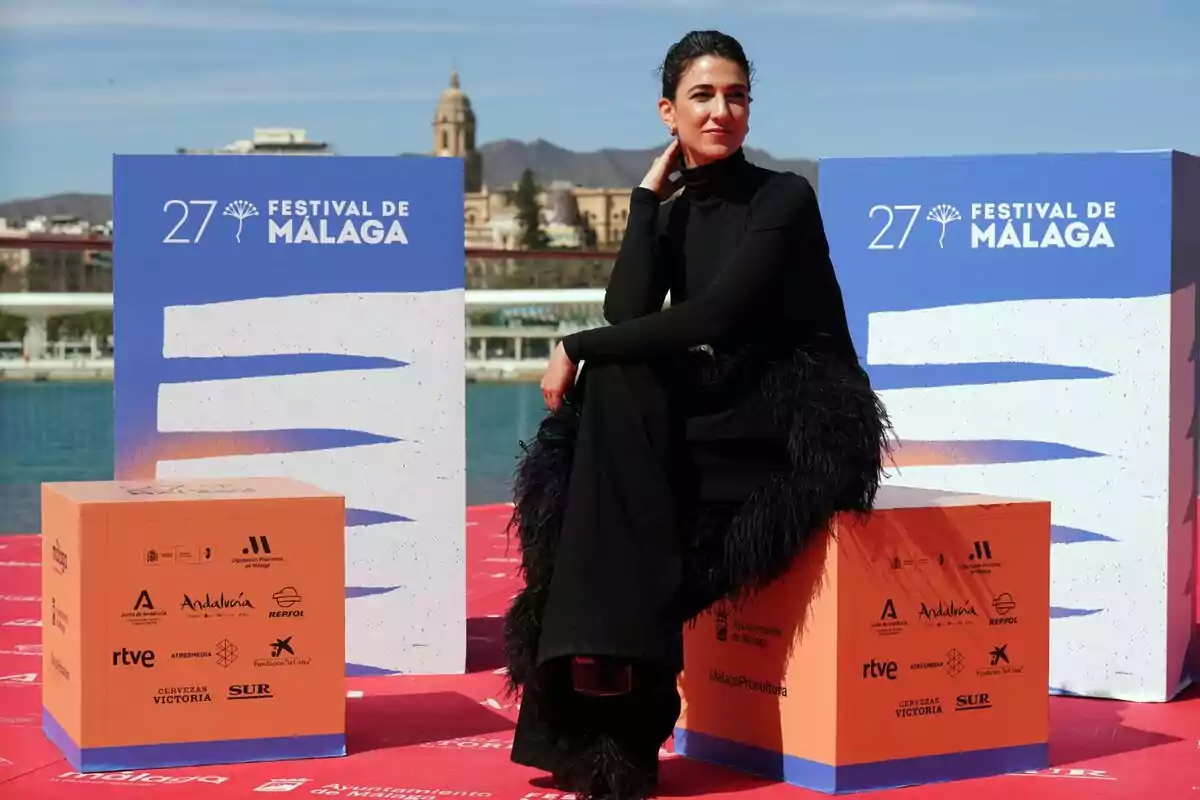 Stéphanie Magnin vestida de negro posa sentada en un cubo naranja durante el 27º Festival de Málaga, con el mar y la ciudad de Málaga de fondo.