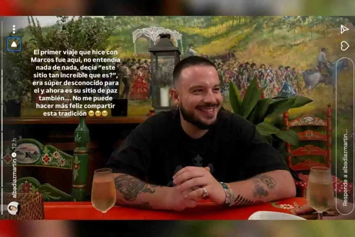 Hombre sonriente sentado en una mesa decorada con copas y sillas coloridas, al fondo un mural pintado con una escena festiva y plantas decorativas Hombre sonriente sentado en una mesa decorada con copas y sillas coloridas, al fondo un mural pintado con una escena festiva y plantas decorativas