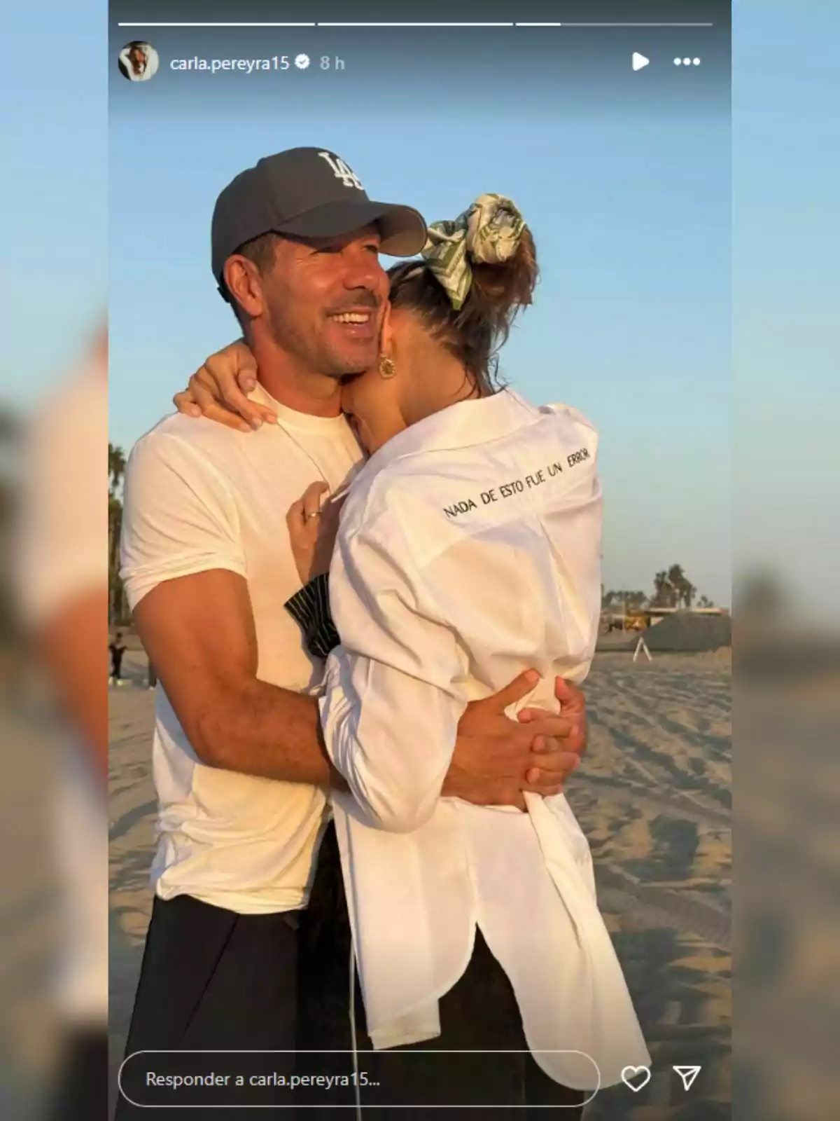 Diego Simeone and Carla Pereyra hugging on the beach at sunset, both smiling, and the woman wearing a white jersey with the phrase Diego Simeone and Carla Pereyra hugging on the beach at sunset, both smiling, and the woman wearing a white jersey with the phrase