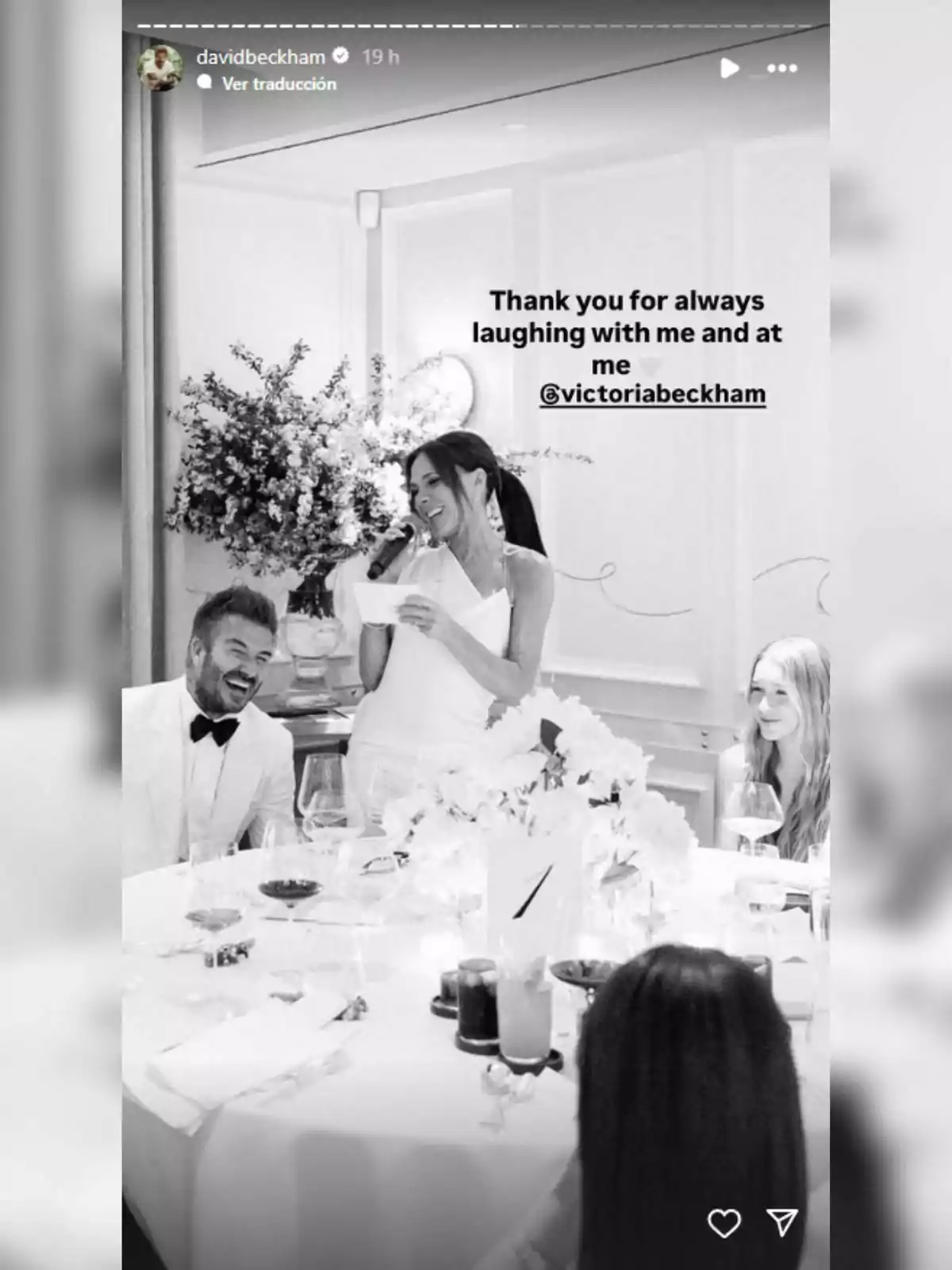 Victoria Beckham sonríe mientras sostiene un micrófono en una cena elegante, acompañada de personas que también se ríen. Victoria Beckham sonríe mientras sostiene un micrófono en una cena elegante, acompañada de personas que también se ríen.