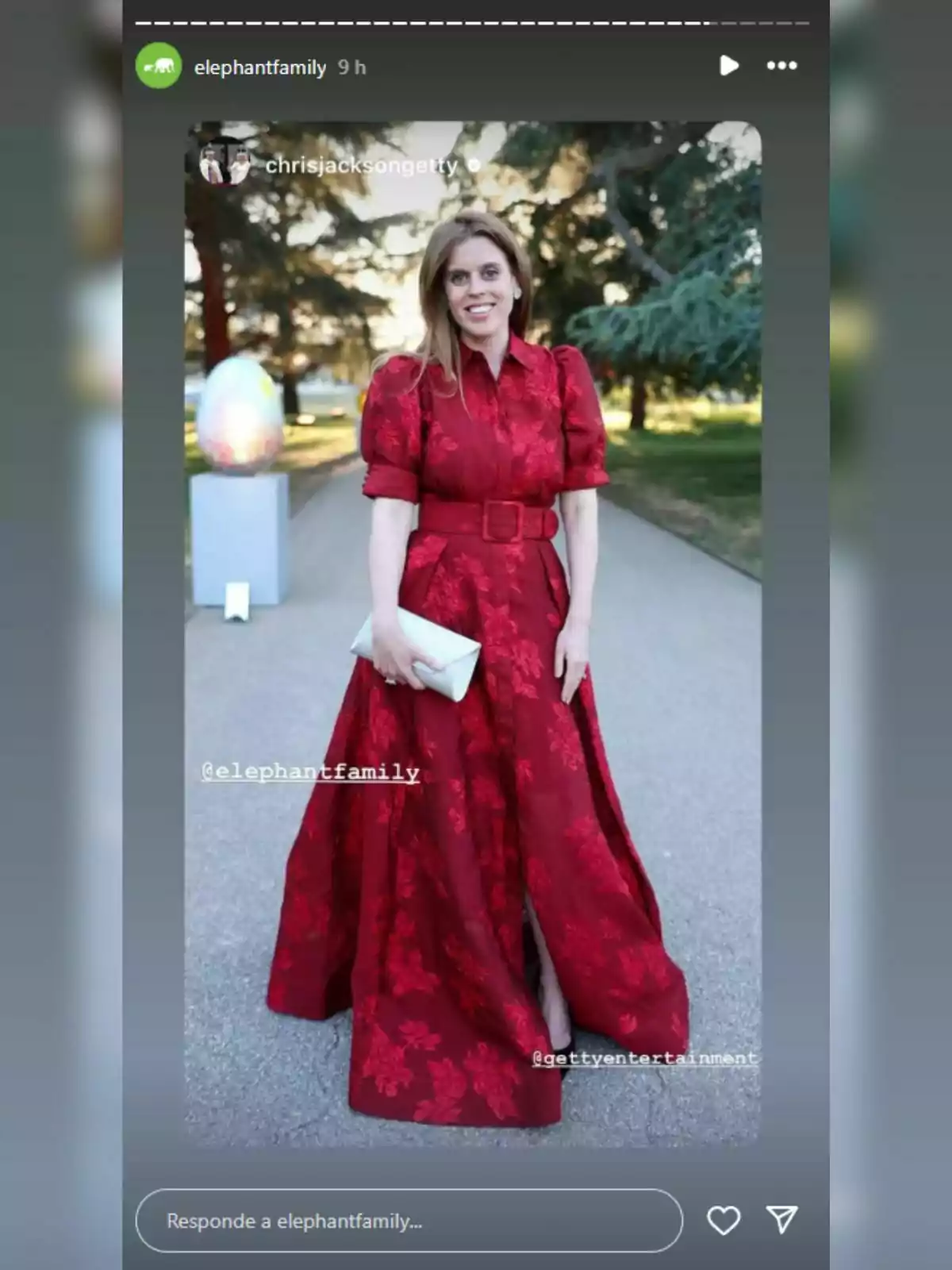 Beatriz of York in an elegant red dress poses outdoors, holding a white handbag, with trees and a sculpture in the background. Beatriz of York in an elegant red dress poses outdoors, holding a white handbag, with trees and a sculpture in the background.