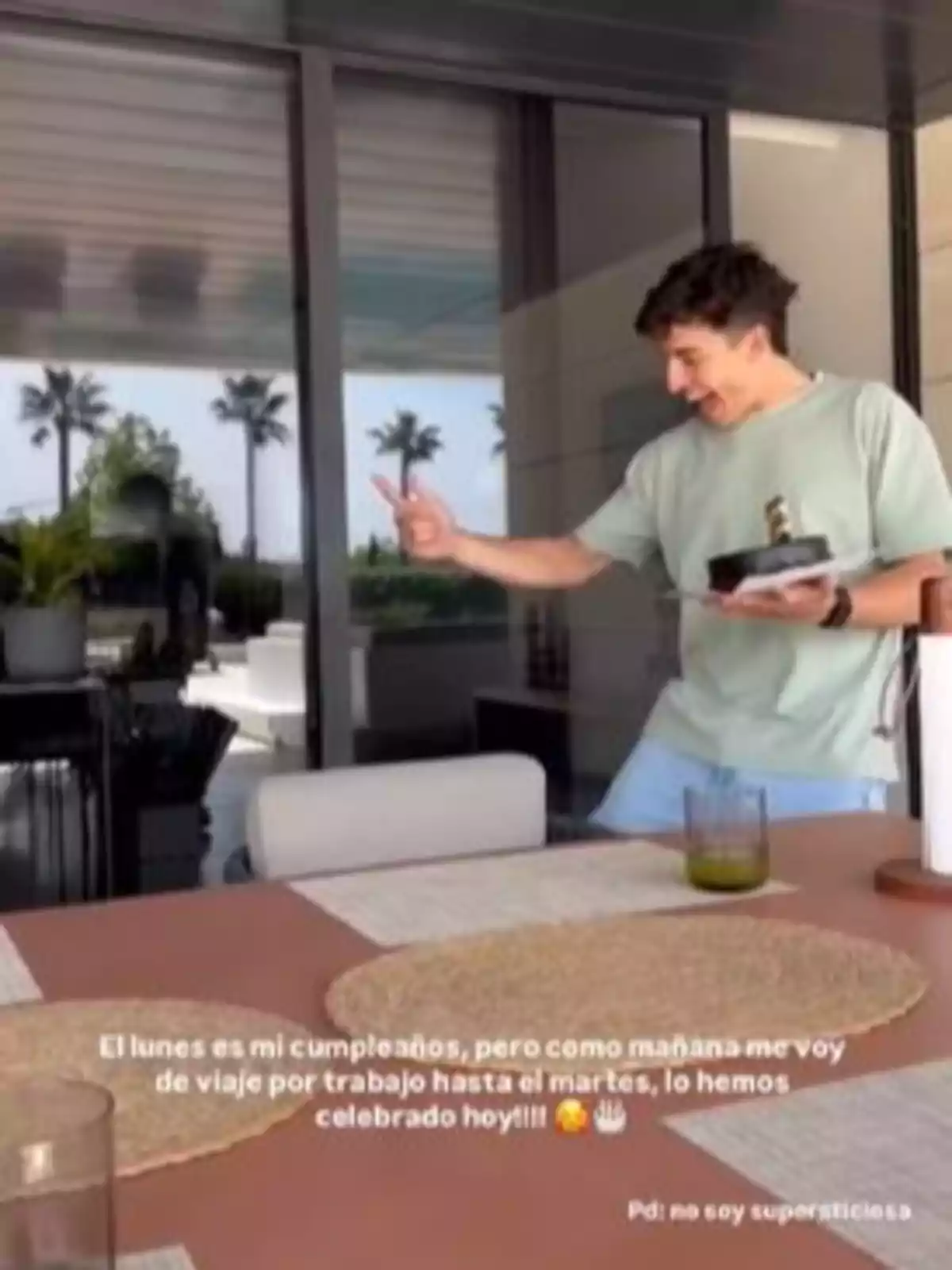 Marc Márquez sonriente sostiene un pastel pequeño con una vela encendida mientras está de pie en una terraza moderna con palmeras al fondo. En la mesa hay un vaso y manteles individuales. Marc Márquez sonriente sostiene un pastel pequeño con una vela encendida mientras está de pie en una terraza moderna con palmeras al fondo. En la mesa hay un vaso y manteles individuales.