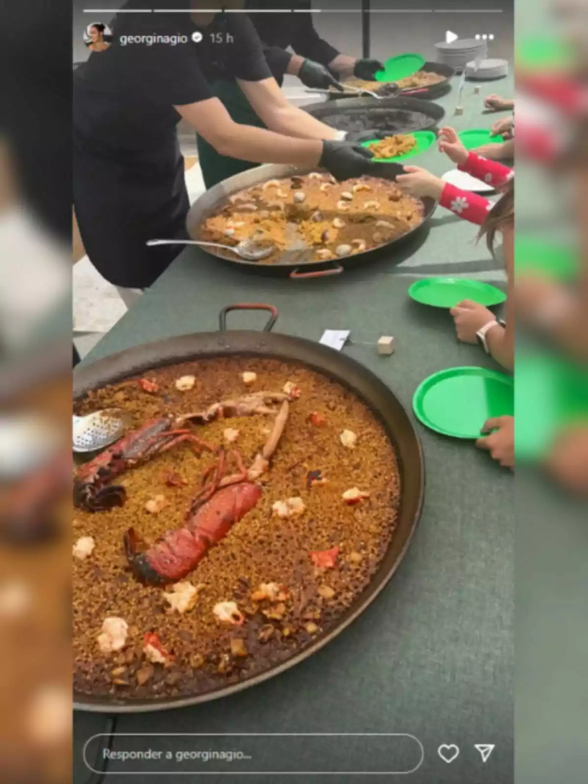 Several people serve seafood rice from large paella pans on a table while children wait with green plates. Several people serve seafood rice from large paella pans on a table while children wait with green plates.