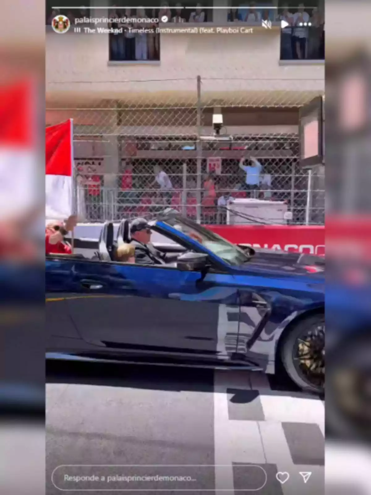 Un coche descapotable azul oscuro circula por una calle con una bandera roja y blanca ondeando, mientras varias personas observan desde detrás de una valla y desde un edificio cercano. Un coche descapotable azul oscuro circula por una calle con una bandera roja y blanca ondeando, mientras varias personas observan desde detrás de una valla y desde un edificio cercano.