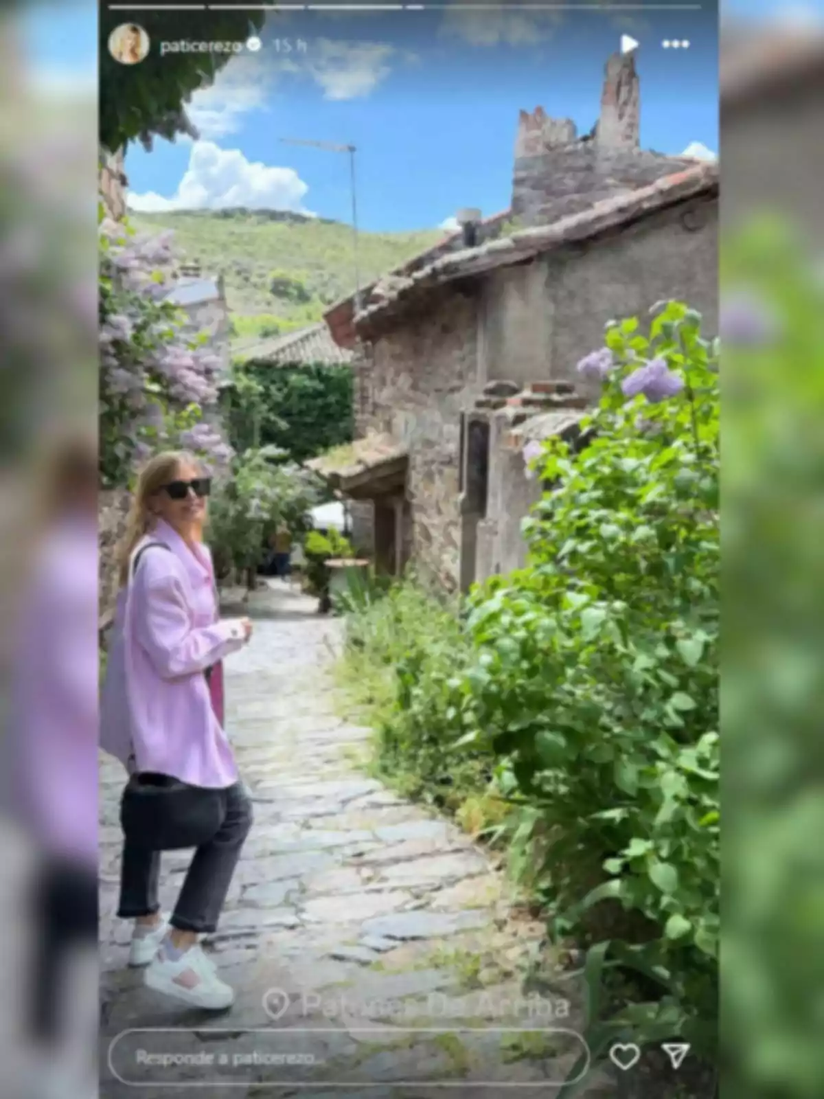 Patricia Cerezo con gafas de sol y chaqueta lila camina por un pintoresco camino empedrado rodeado de vegetación y casas de piedra, con un cielo azul y montañas al fondo. Patricia Cerezo con gafas de sol y chaqueta lila camina por un pintoresco camino empedrado rodeado de vegetación y casas de piedra, con un cielo azul y montañas al fondo.